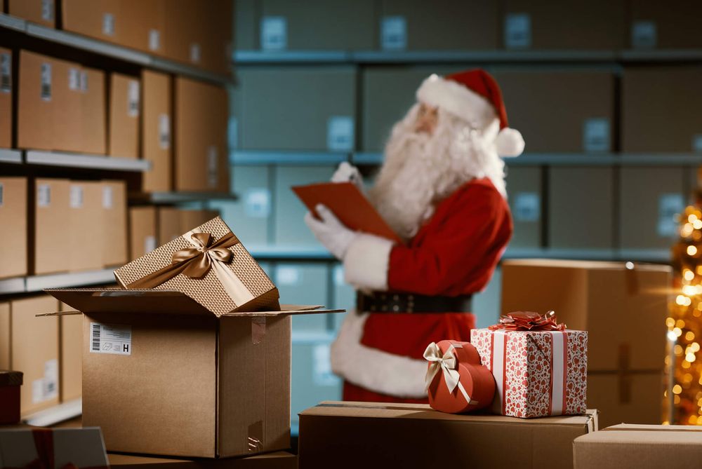 Père Noël vérifiant une liste dans un entrepôt rempli de colis, avec un carton ouvert, des cadeaux emballés et un sapin illuminé.