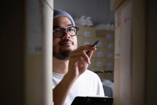 Pessoa com uma prancheta a inventariar caixas de expedição etiquetadas num espaço de preparação de encomendas